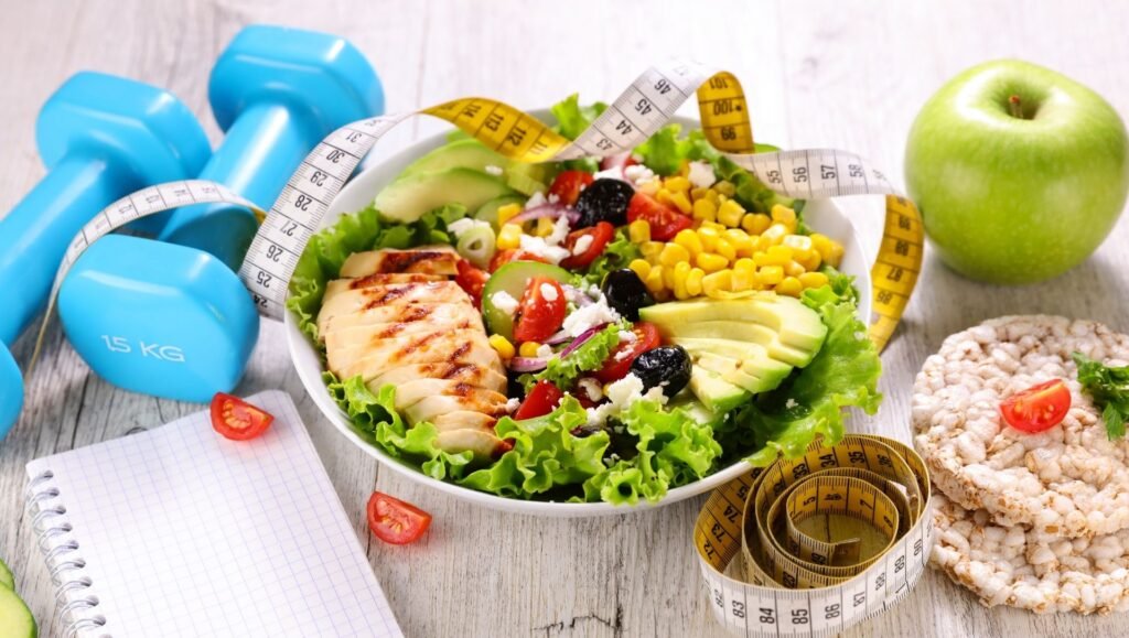 Healthy salad bowl with grilled chicken, vegetables, avocado, measuring tape, dumbbells, apple, and rice cakes.