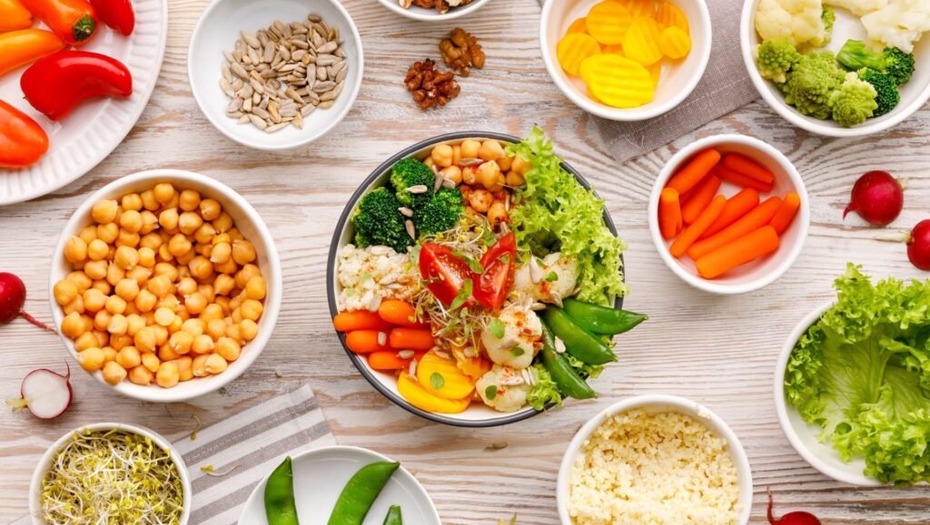Colourful plant-based salad bowl surrounded by fresh vegetables, chickpeas, nuts, seeds, and leafy greens.