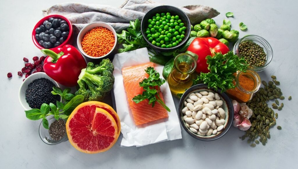 Assortment of healthy whole foods including salmon, legumes, fresh vegetables, fruits, herbs, seeds, and olive oil on a light background.