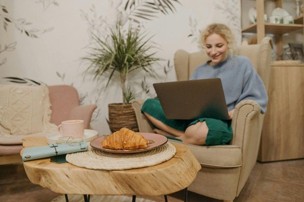 A woman sitting cross-legged on a beige armchair, working on a laptop in a cozy living room with plants. A wooden coffee table in the foreground holds a croissant on a plate, a cup of coffee, a notebook, and glasses.