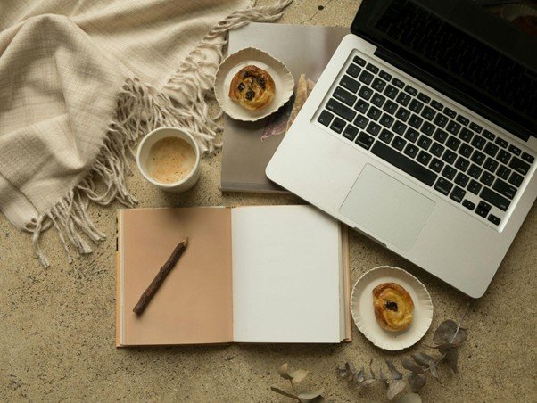 A cozy workspace setup with an open laptop, a blank notebook, a cup of coffee, and pastries on a textured surface beside a beige fringed blanket.