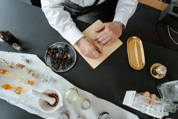 Person writing in a notebook at a dark table with glass bottles, dried herbs, and golden containers arranged neatly around them.