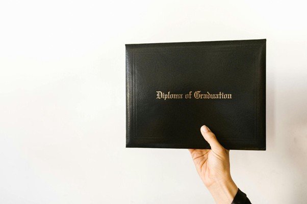 A hand holds up a black diploma cover with the words "Diploma of Graduation" embossed in gold on the front against a plain white background.