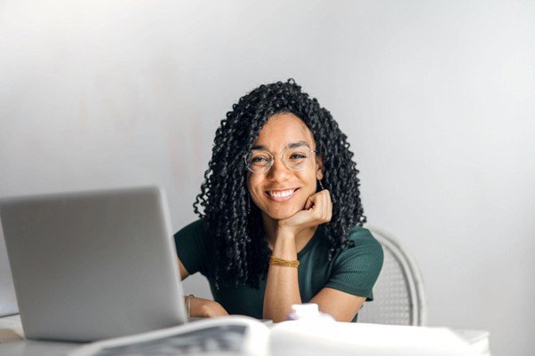 A smiling young woman with dark, braided hair and glasses sits happily at a desk with an open laptop and books in front of her.