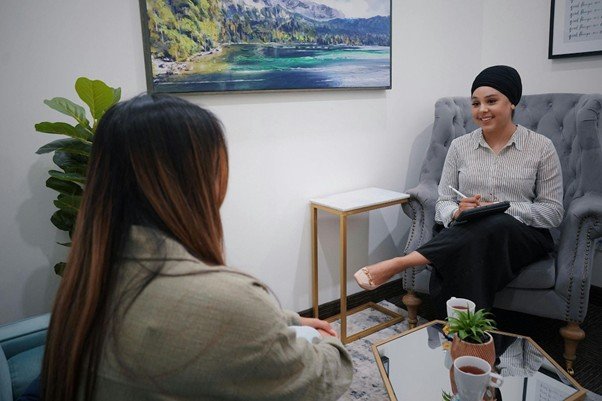 A therapist, wearing a headscarf, smiles while sitting in a large armchair and speaking with a client in a modern, comfortable office setting.