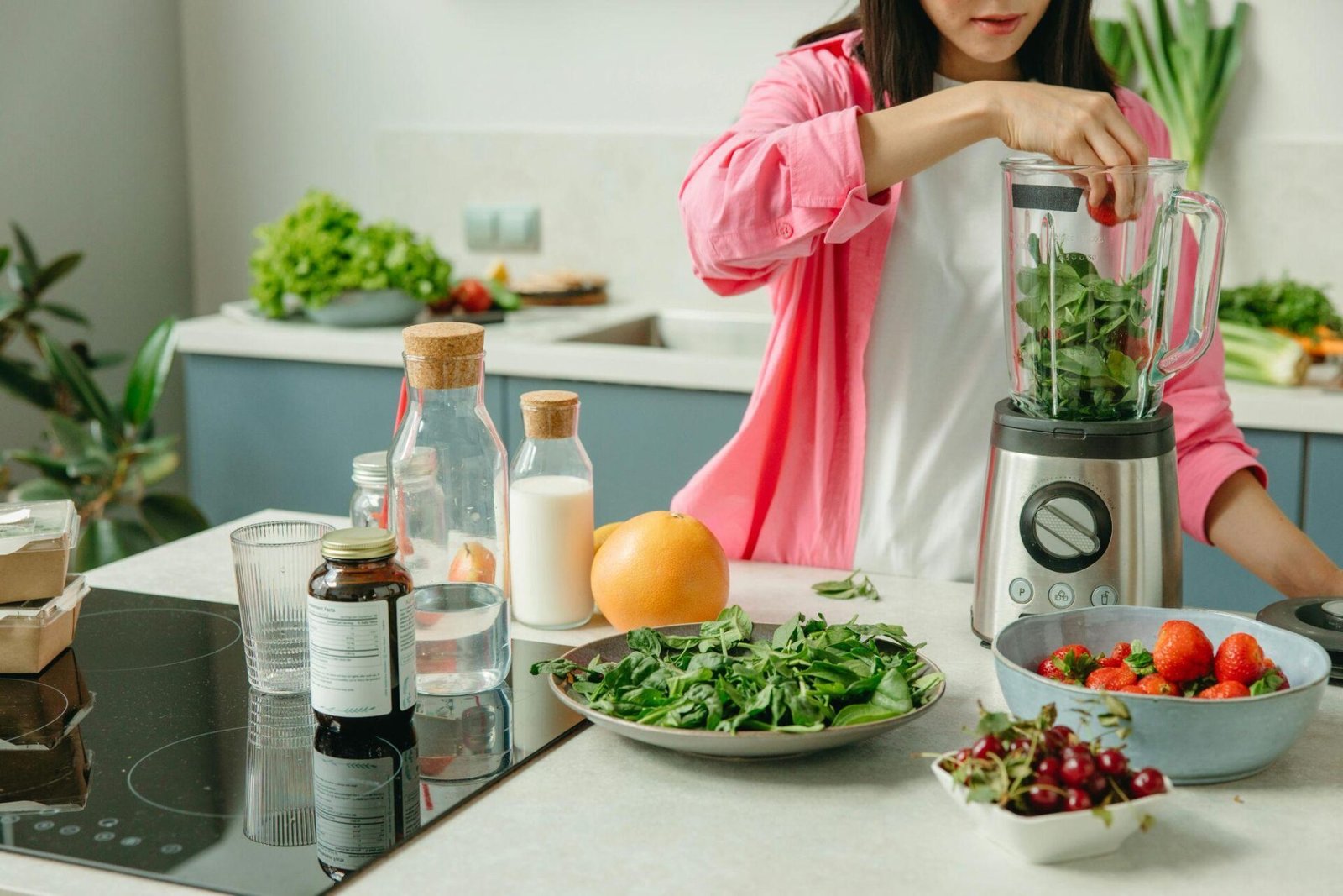 A high-angle shot showing a fit woman with her hair pulled back in a ponytail, crouching in front of a laptop on a low wooden coffee table. She is wearing a black tank top and shorts. On the table, there is a plate with bananas, green apples, and broccoli, a handwritten note, a glass of water, and a dark water bottle. The background shows a living room with a black leather sofa, a patterned rug, and concrete-style flooring, suggesting she is preparing for a workout or reviewing nutrition/fitness plans.